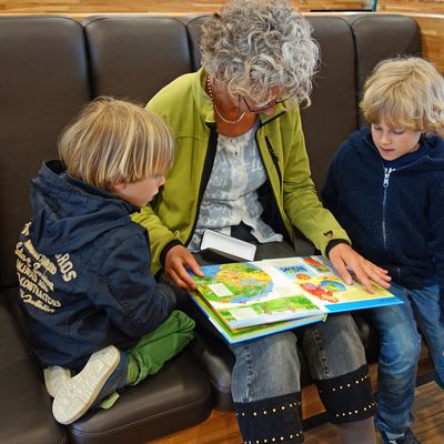 Eine Frau sitzt zwischen zwei Kindern. Alle drei Personen schauen in ein Kinderbuch, dass die Frau aufgeschlagen auf ihrem Scho platziert hat.